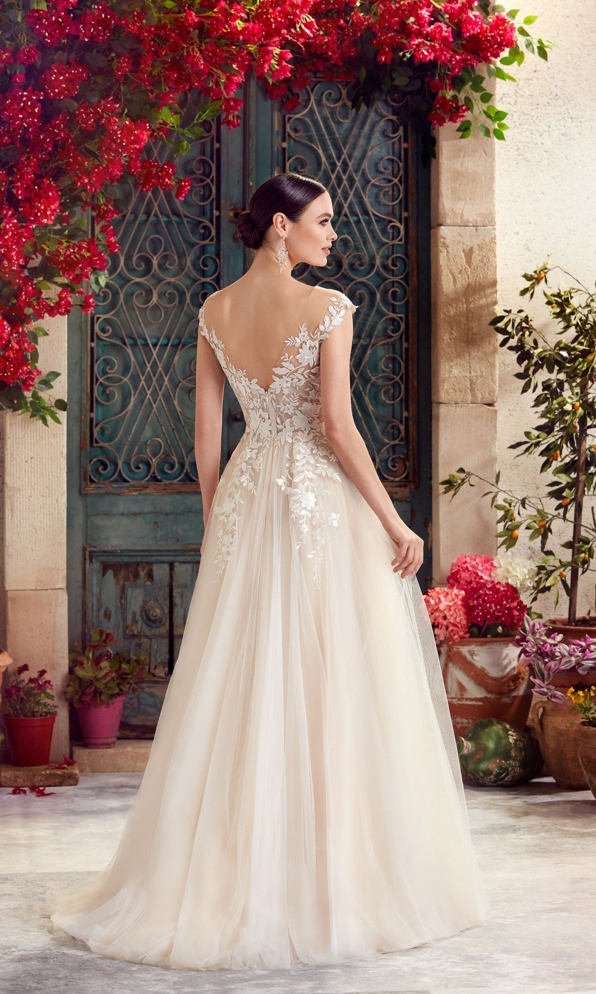 A woman in the Alyce 5090 long formal dress, featuring white lace embroidery and an elegant a-line tulle skirt, stands outdoors facing away near a teal door framed by vibrant red flowers and potted plants.
